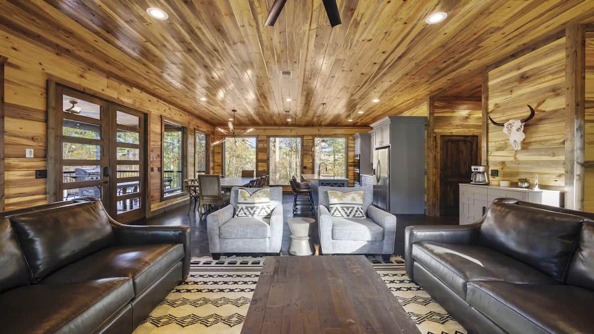 Interior de una cabaña con paredes de madera, una zona de estar central con sofás y sillones, una mesa de comedor y una cocina.