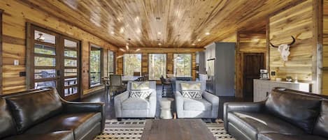 Interior de una cabaña con paredes de madera, una zona de estar central con sofás y sillones, una mesa de comedor y una cocina.