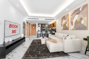 A modern living room with a sofa, a coffee table, and a TV. There is a kitchen area with a refrigerator and a microwave. The room has a marble floor and a black rug. - Stayshort - Spacious 1 Bedroom Apartment in JVT for up to 4 Guests (Dubai)