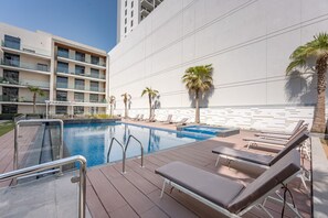 A swimming pool with lounge chairs and palm trees, surrounded by a modern building with balconies. - Stayshort - Spacious 1 Bedroom Apartment in JVT for up to 4 Guests (Dubai)