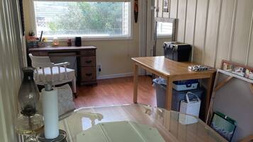 A room with a glass-top table, white chairs, a wooden desk, and a window with blinds.