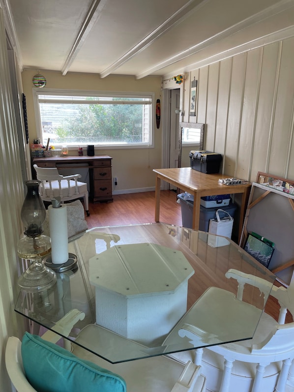 A room with a glass-top table, white chairs, a wooden desk, and a window with blinds.