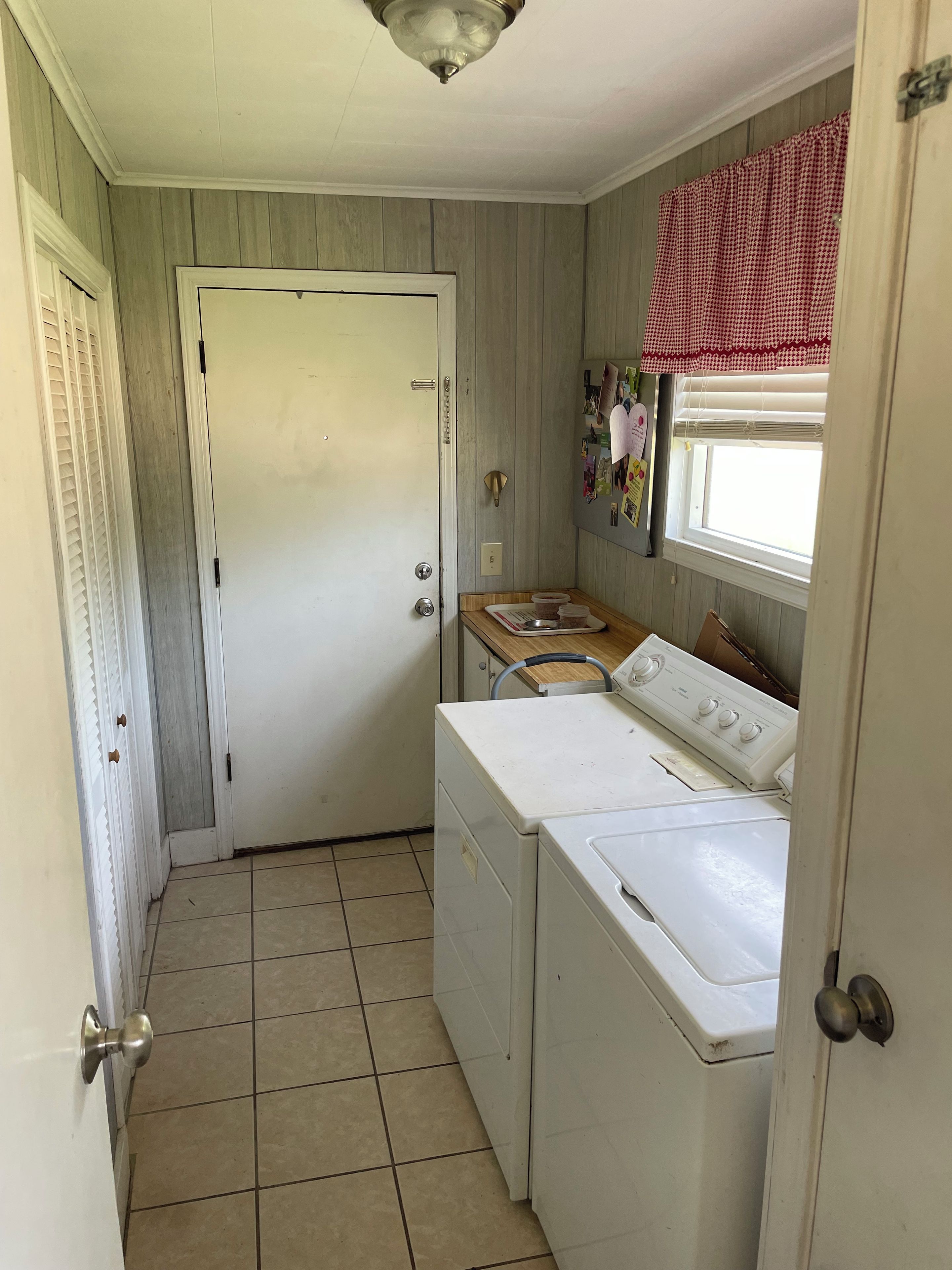 A laundry room with a white door, a washing machine, a dryer, and a window with a red checkered curtain.