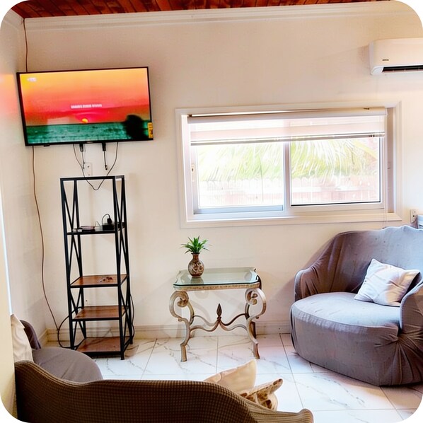 A living room with a television, a window with a view of greenery, a sofa, and a decorative table.