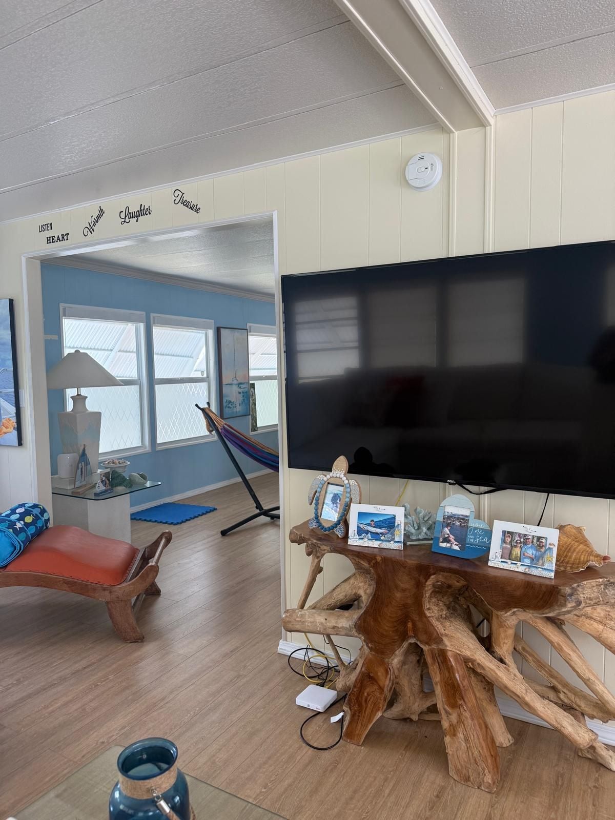 Een kamer met een grote flatscreen televisie op een houten standaard, een hangmat, een rode, beklede bank en een muur met motiverende woorden.