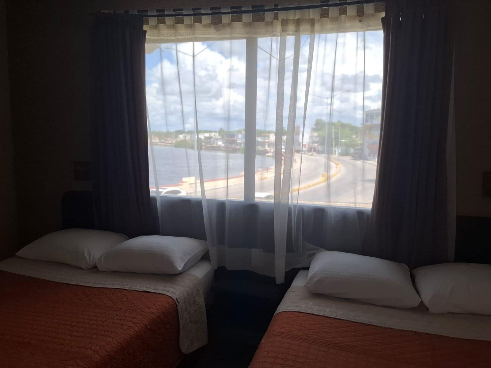 Habitación de hotel con dos camas, vista a una carretera y al agua, y una ventana con cortinas traslúcidas.
