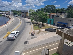 A street view with a white car, a motorbike, and a parked car. There are buildings, trees, and a clear sky.