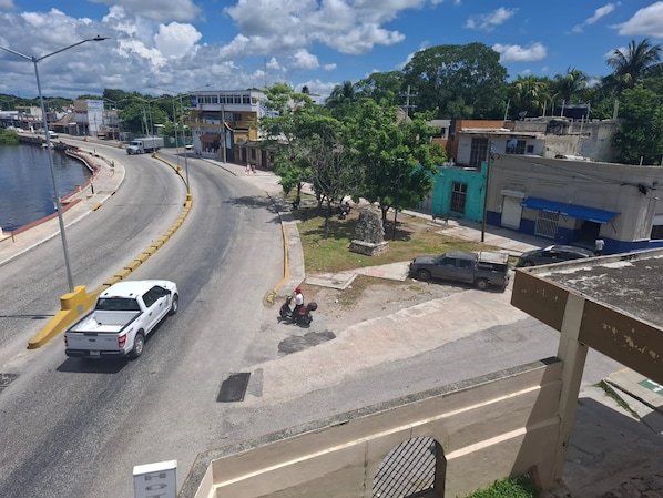 A street view with a white car, a motorbike, and a parked car. There are buildings, trees, and a clear sky.