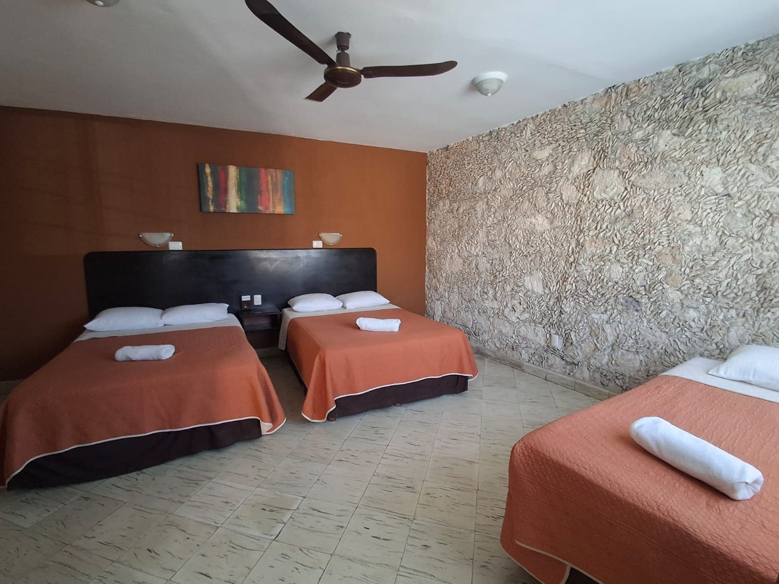 Habitación de hotel con dos camas, ventilador de techo, pared con textura de piedra y un cuadro.