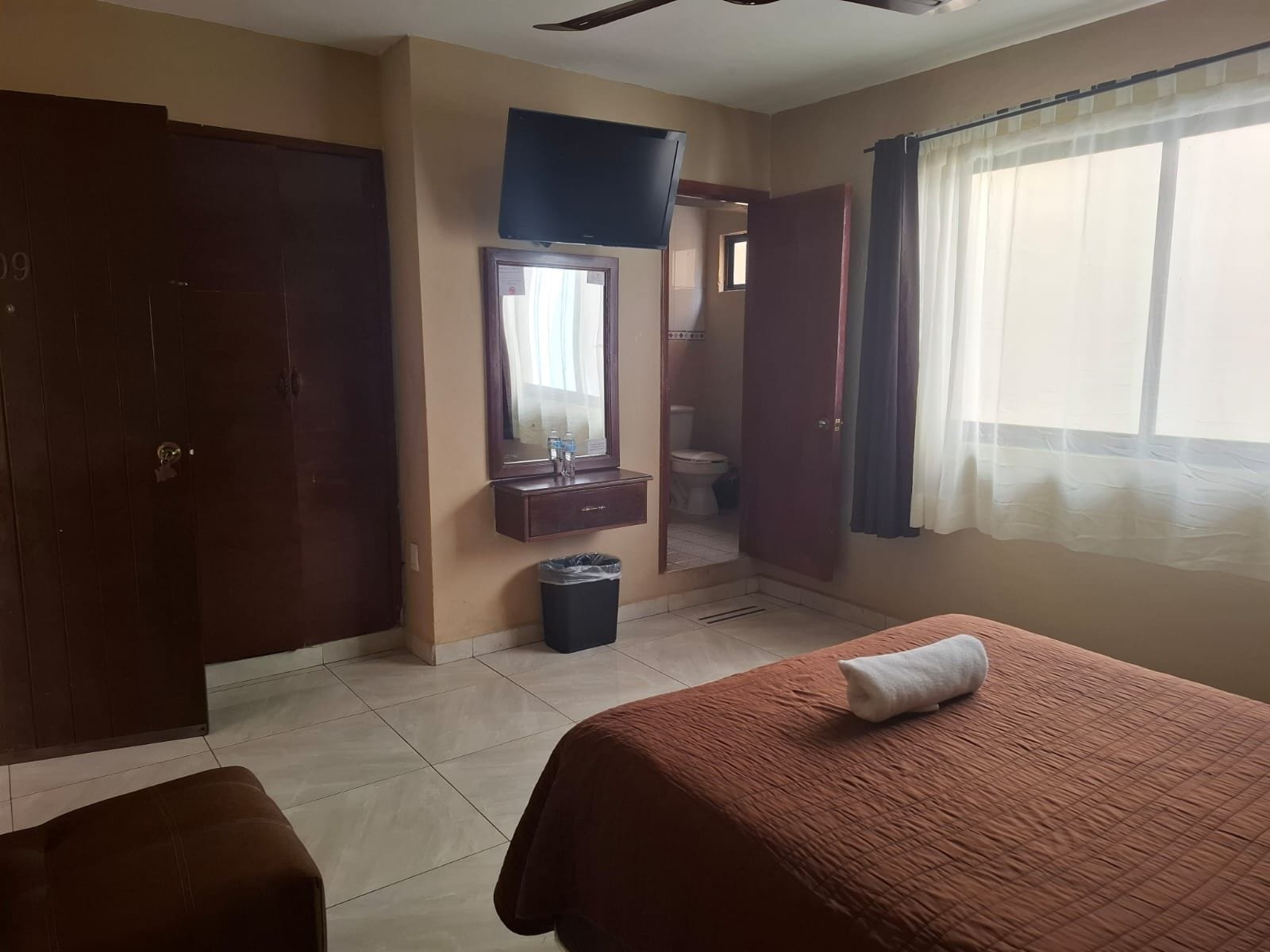 Habitación de hotel con cama, una mesita, espejo, televisión, ventana con cortinas y puerta.