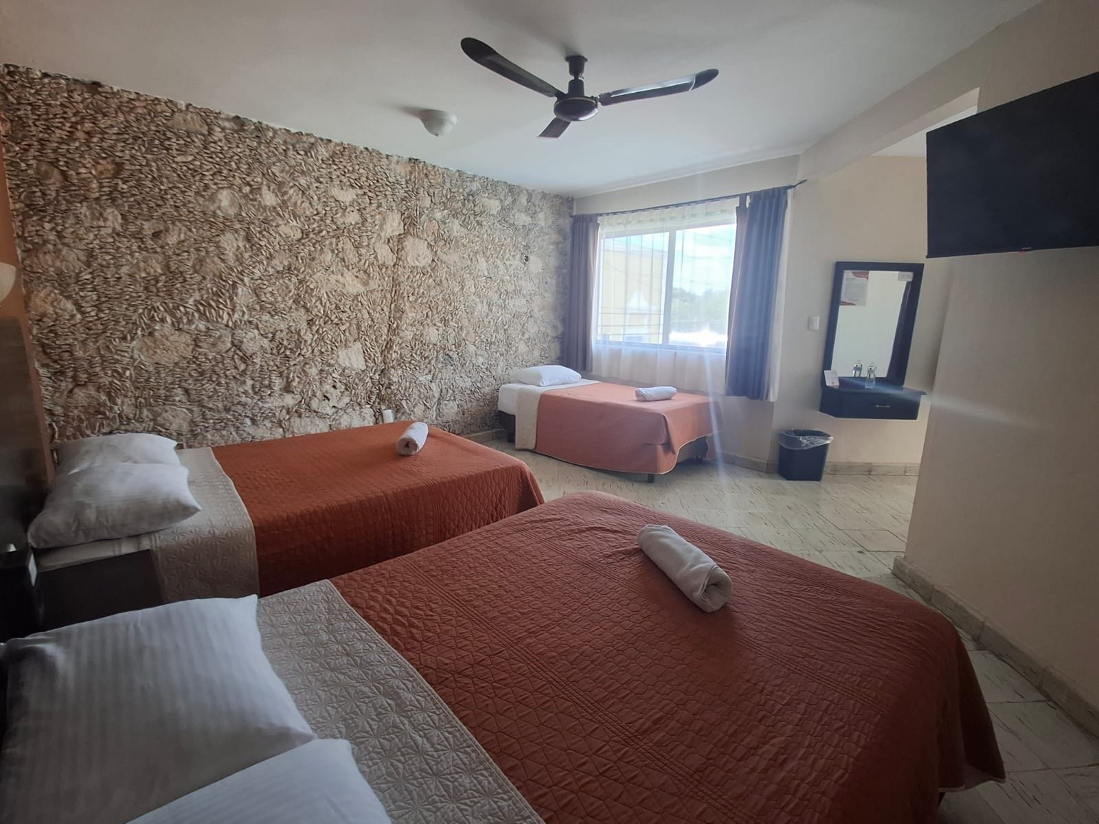 Habitación de hotel con dos camas, una pared de piedra, ventilador de techo, ventana con cortinas y un lavamanos.