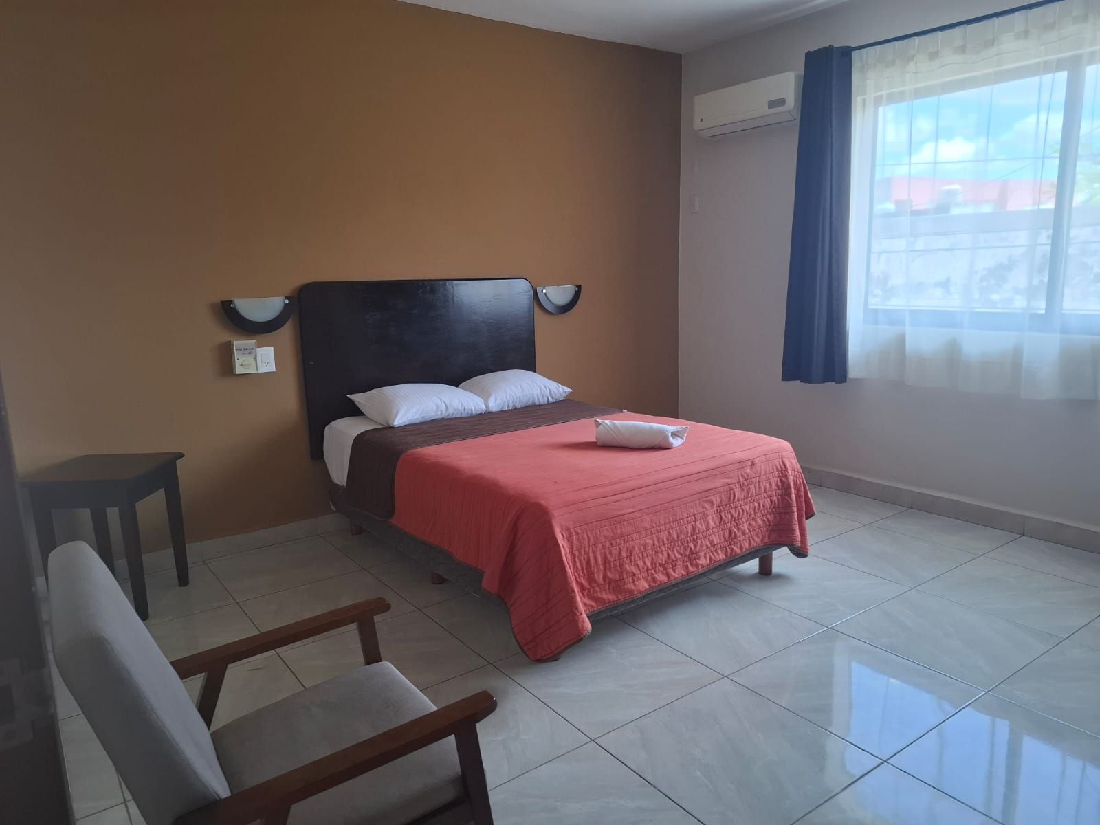 Una habitación de hotel con una cama, una silla, una mesita de noche y un ventanal con cortinas.