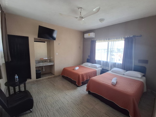 A hotel room with two beds, a desk, a chair, a television, an air conditioning unit, and a window with curtains.