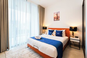 A hotel room with a bed, two bedside lamps, a nightstand, a wall clock, and a window with curtains. - Rentico Lux 1BR Burj View Paragon (Dubai)