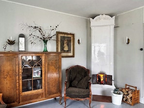 A room with a wooden cabinet, a chair, a fireplace, and a vase with branches. - Farmhouse with tiled stoves near lake and forest (Asarum)