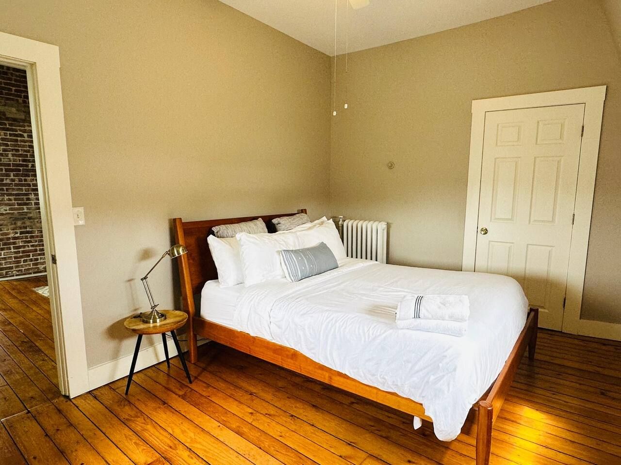 Une chambre à coucher avec un lit en bois, une literie blanche, une petite table ronde avec une lampe et un radiateur.