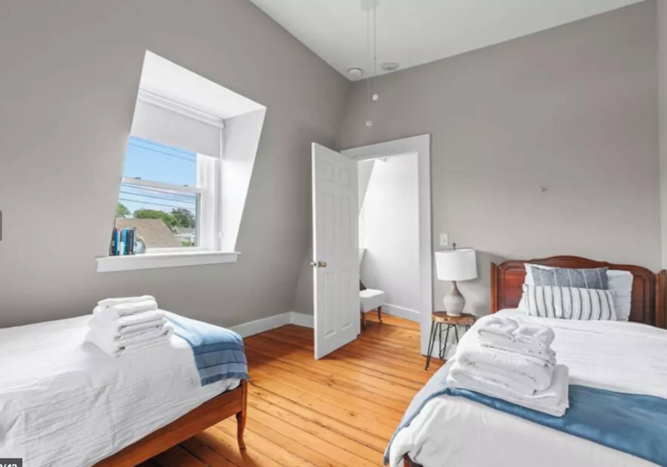 A bedroom with two beds, a window, a door, and a bedside table with a lamp.