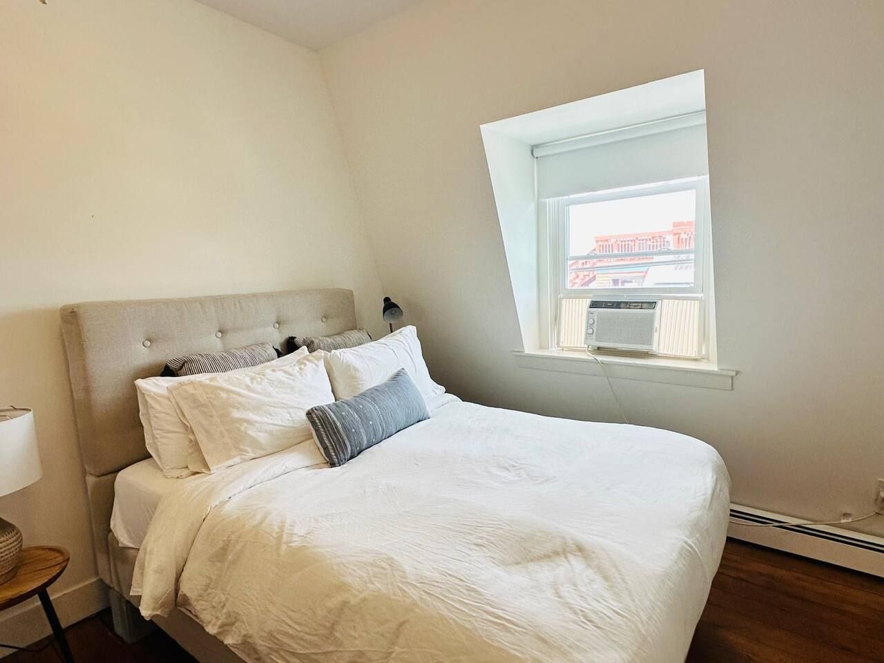 A bedroom with a bed, a nightstand, a lamp, a window with a view, and a thermostat.