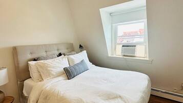 A bedroom with a bed, a nightstand, a lamp, a window with a view, and a thermostat.