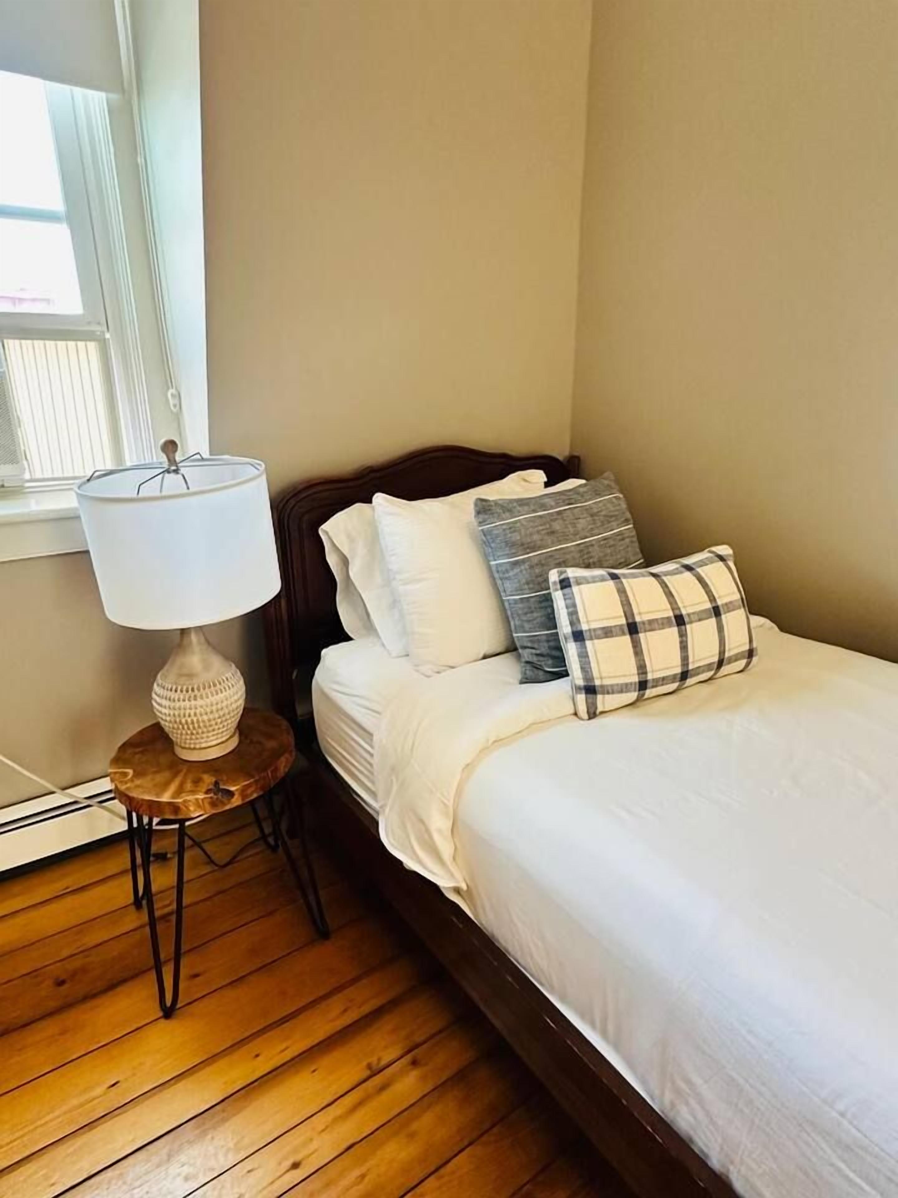 A bedroom with a wooden floor, a bed with white bedding, a round side table with a lamp, and a window with blinds.