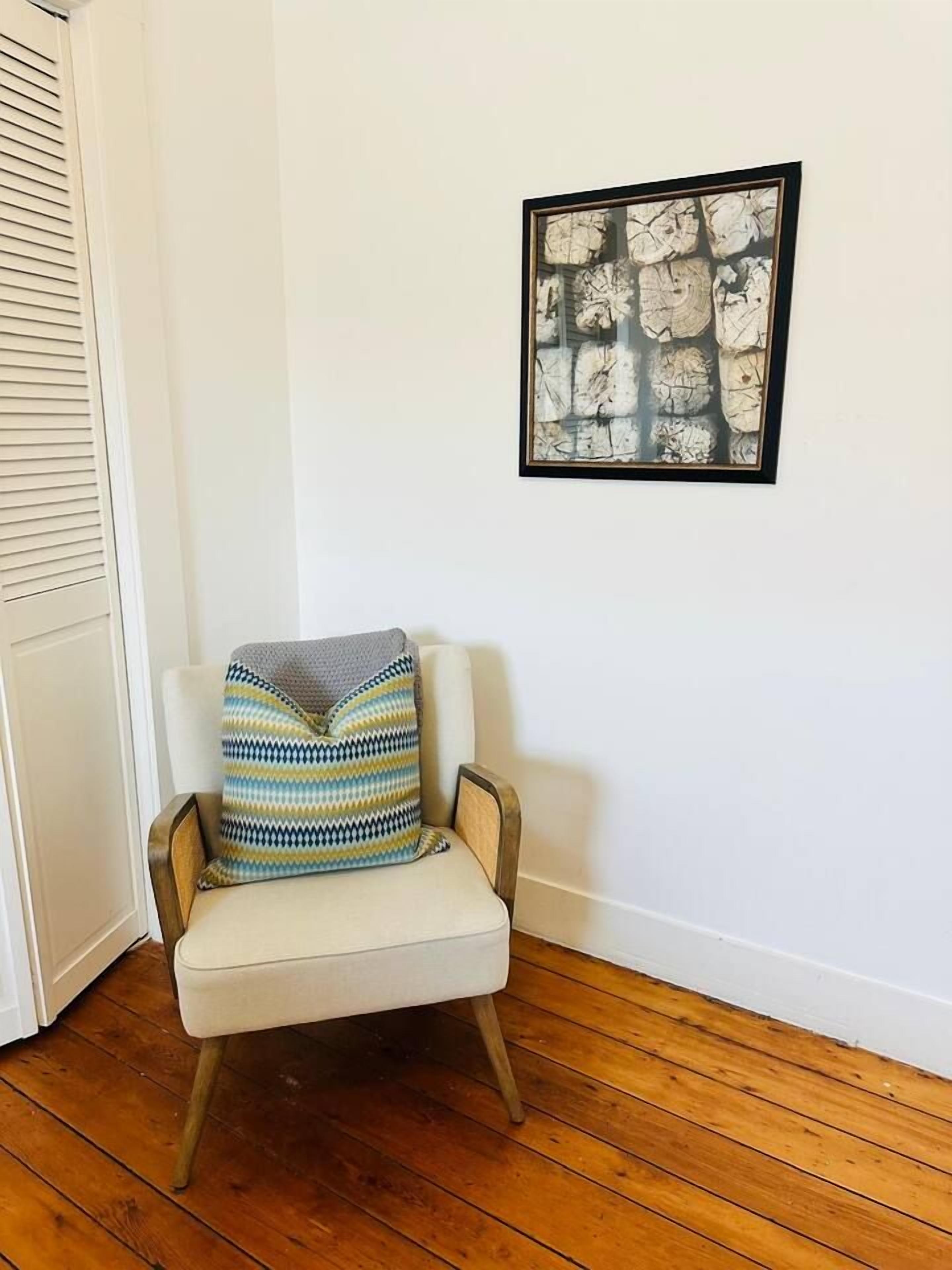 A corner room with a wooden floor, a white wall, a framed abstract artwork, and a chair with a patterned throw pillow.