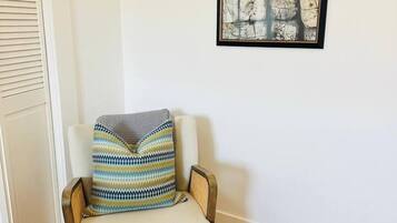 A corner room with a wooden floor, a white wall, a framed abstract artwork, and a chair with a patterned throw pillow.