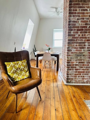 Een kamer met een bakstenen muur, een houten vloer, een bruine leren stoel met een gestreept kussen, een eettafel met stoelen en een raam dat natuurlijk licht binnenlaat.