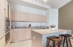 A modern kitchen with light wood cabinets, a central island with stools, a built-in oven, and a coffee maker. - 40th Floor Ocean Penthouses Together (Hollywood)