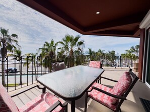 Ein Balkon mit Glastisch und Stühlen, der Blick auf einen Strand mit Palmen und einem klaren Himmel bietet.