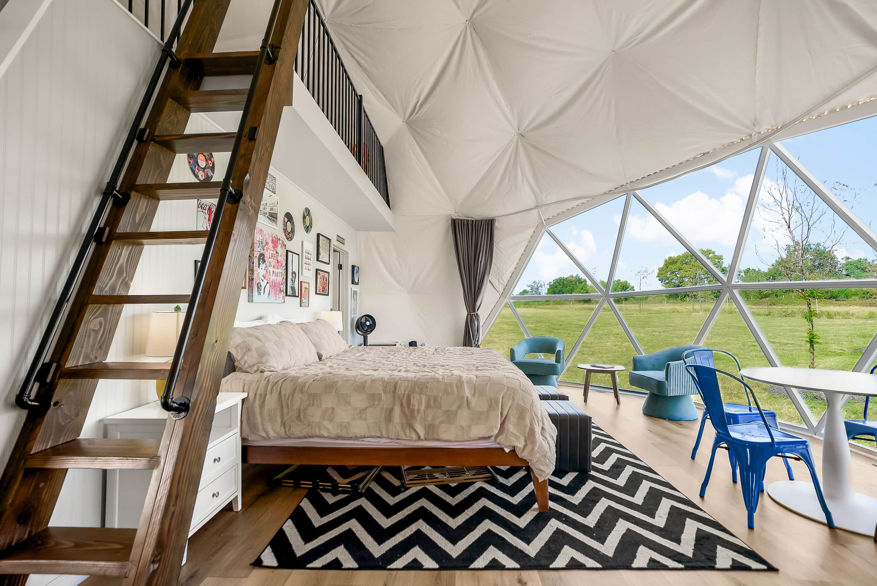 Ein Schlafzimmer mit einem Bett, einer Kommode, einem Stuhl und einem Tisch. Eine Leiter führt zu einem Erker mit einem Fenster.