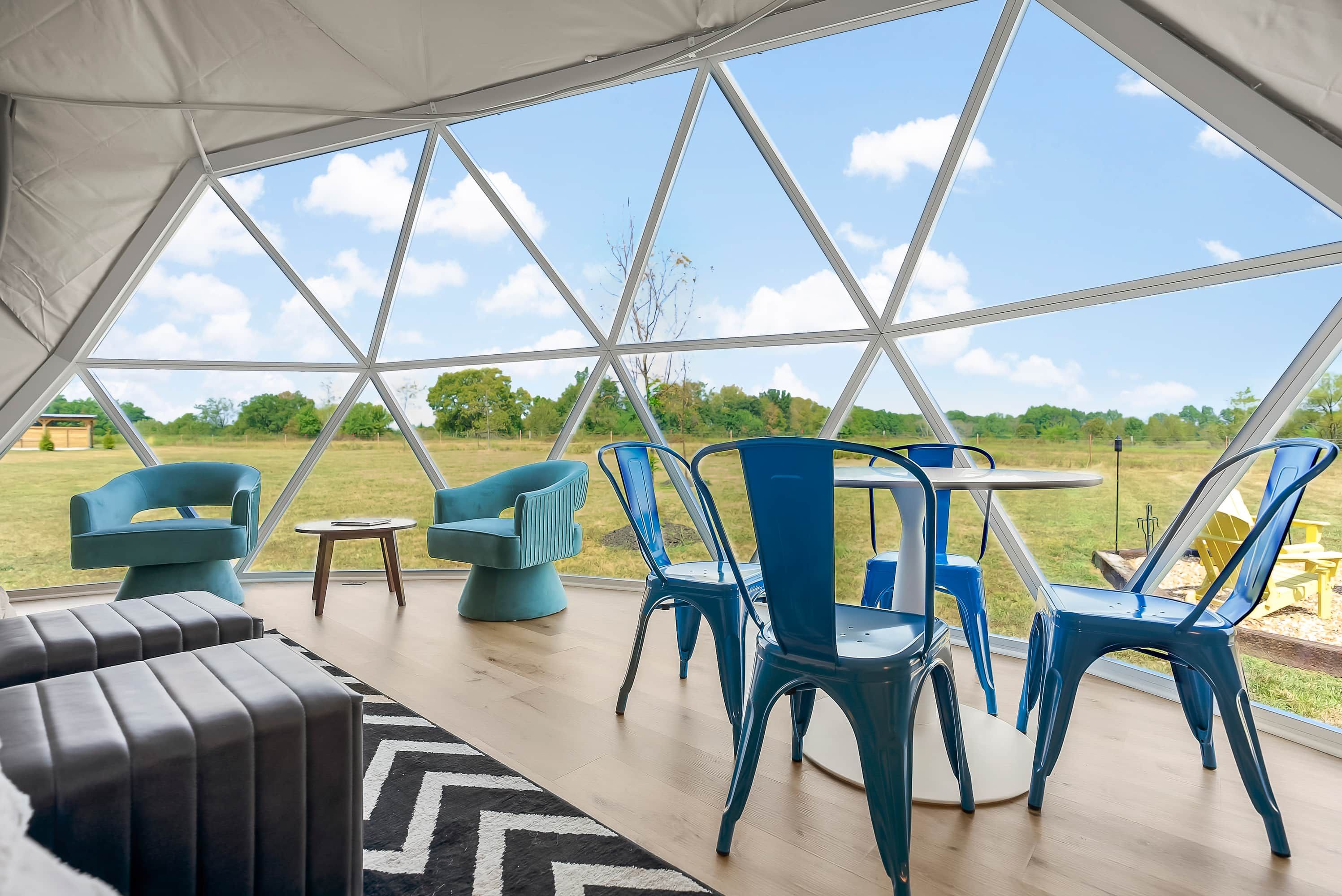 A tent with a geometric roof structure, blue chairs, a round table, and a black and white rug.