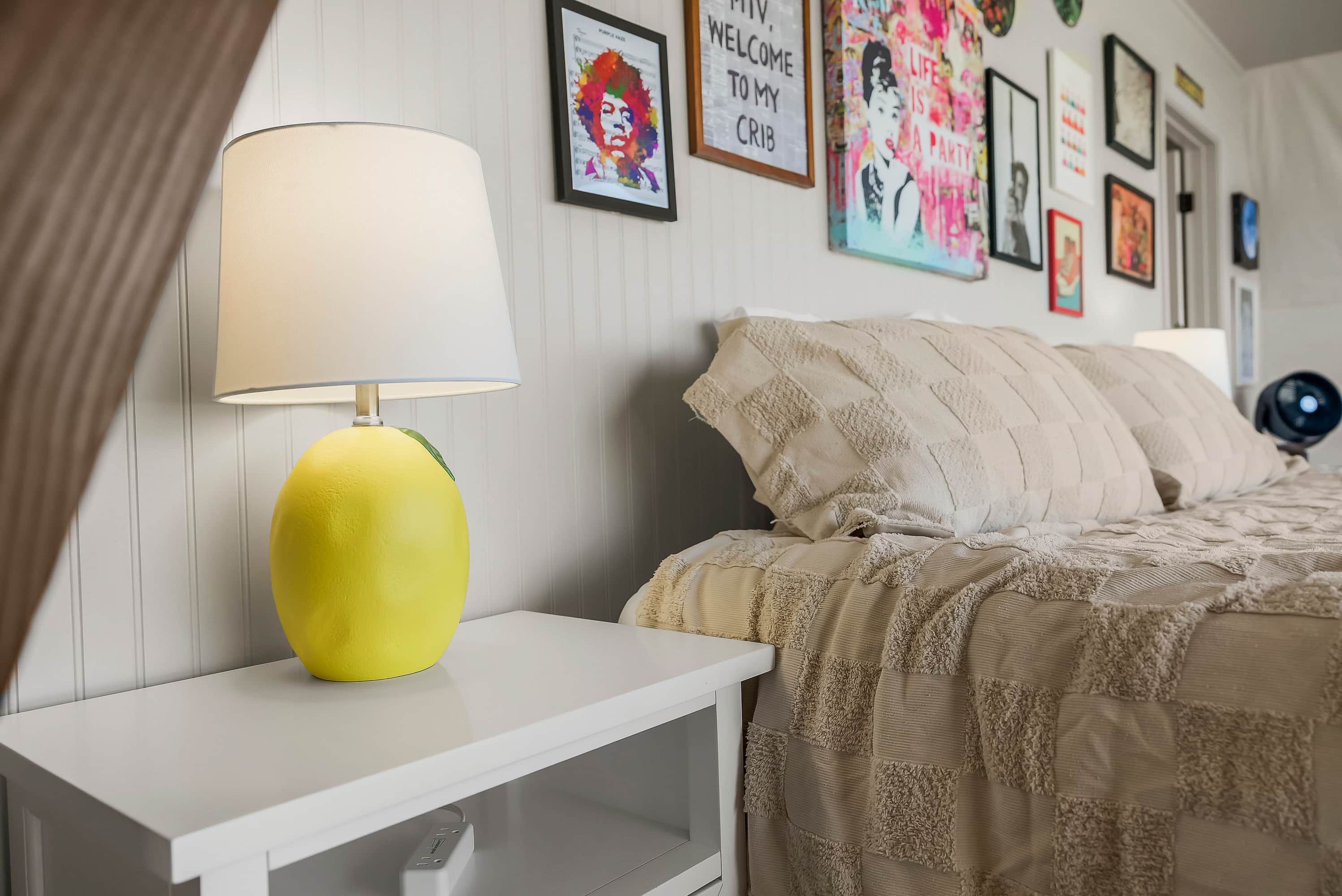Un dormitorio con una mesita de noche blanca, una lámpara amarilla, una cama con una colcha y almohadas color beige, y varias obras de arte enmarcadas en la pared.