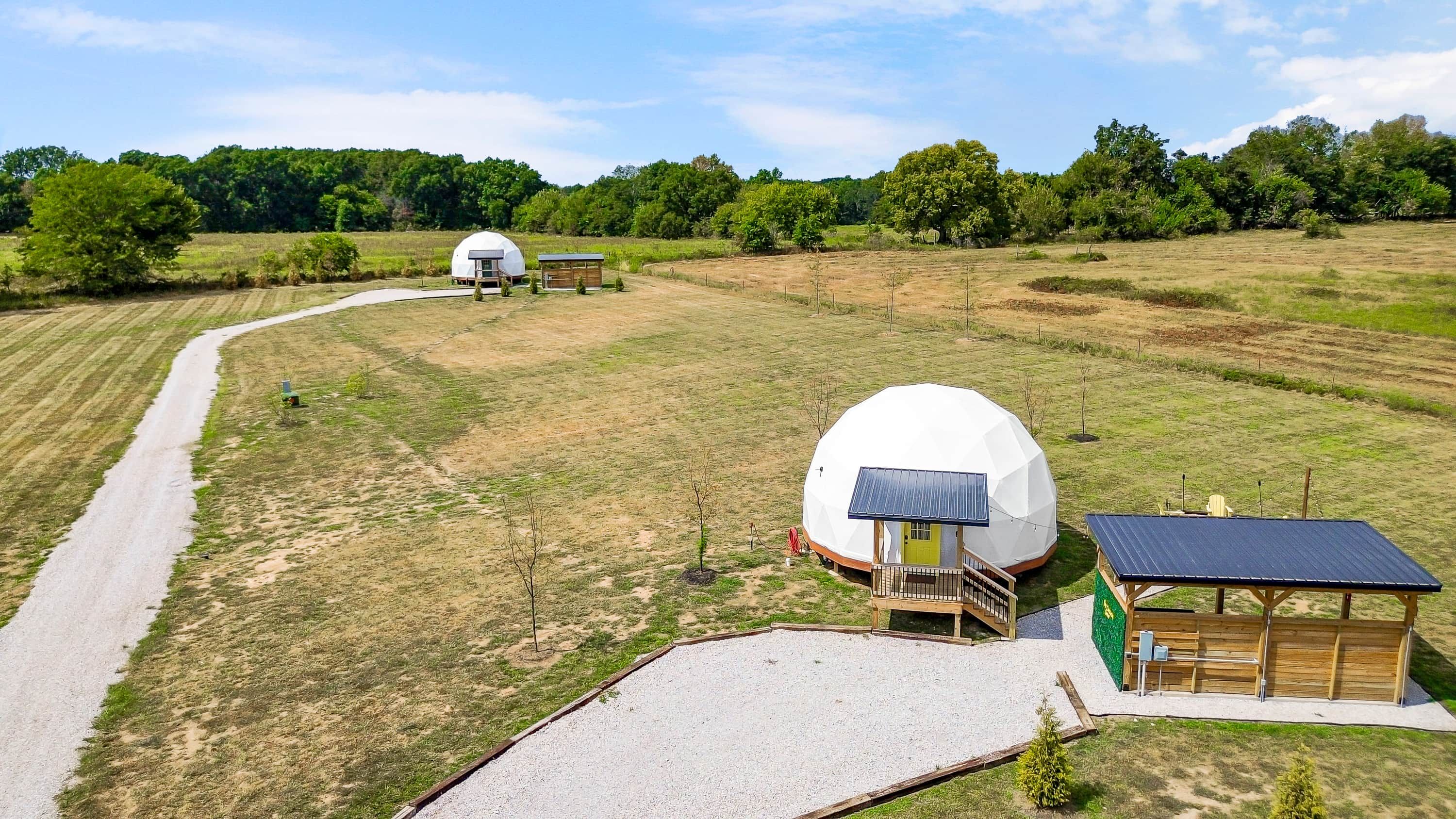 A spherical structure with a wooden deck and a small building with a green roof, surrounded by a gravel path and grassy fields.