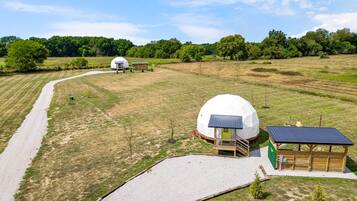 A spherical structure with a wooden deck and a small building with a green roof, surrounded by a gravel path and grassy fields.