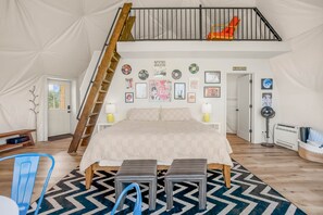 Un dormitorio con un altillo, una cama, un banco, una silla y un ventilador.