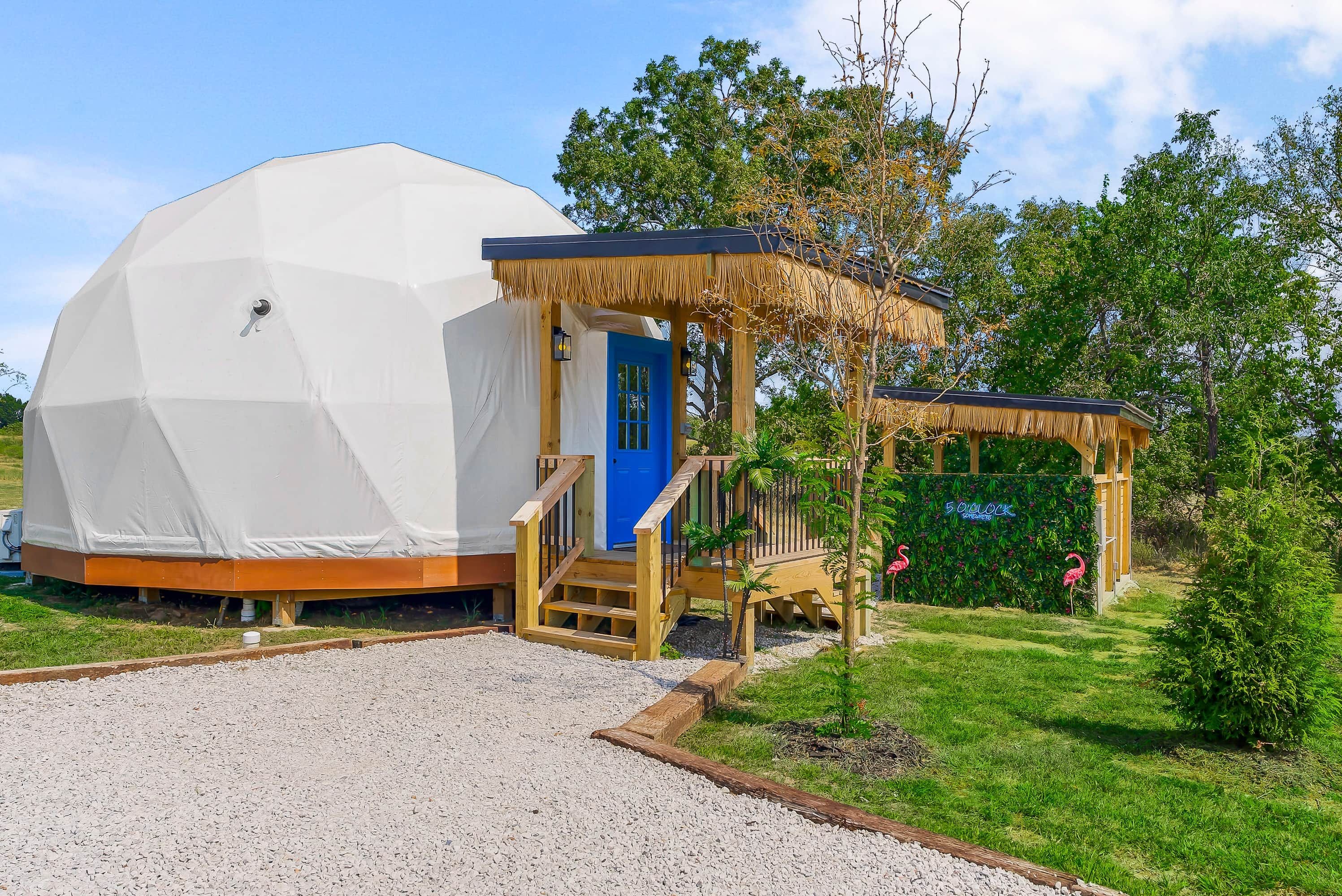 A geodesic dome structure with a wooden porch and a blue door, surrounded by greenery and a clear sky.