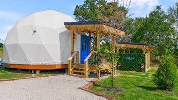 A geodesic dome structure with a wooden porch and a blue door, surrounded by greenery and a clear sky.