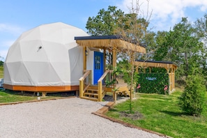 A geodesic dome structure with a wooden porch and a blue door, surrounded by greenery and a clear sky.