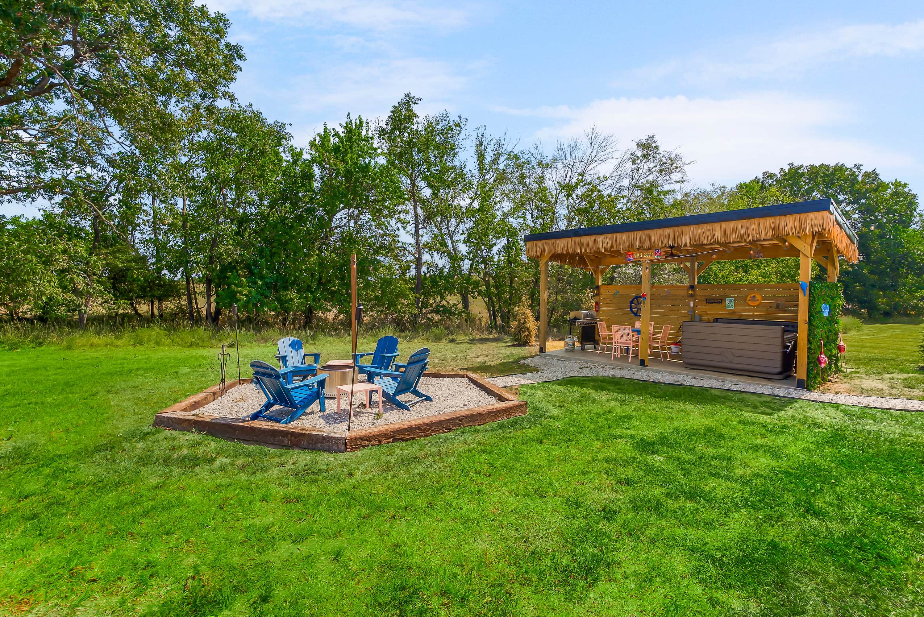 Een prieel met rieten dak, omgeven door een gazon met blauwe Adirondack-stoelen.