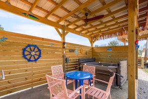 Une terrasse extérieure dotée d’une plateforme en bois, d’une table bleue, de chaises roses, d’un barbecue et d’un plafond en bois avec une section recouverte de chaume.