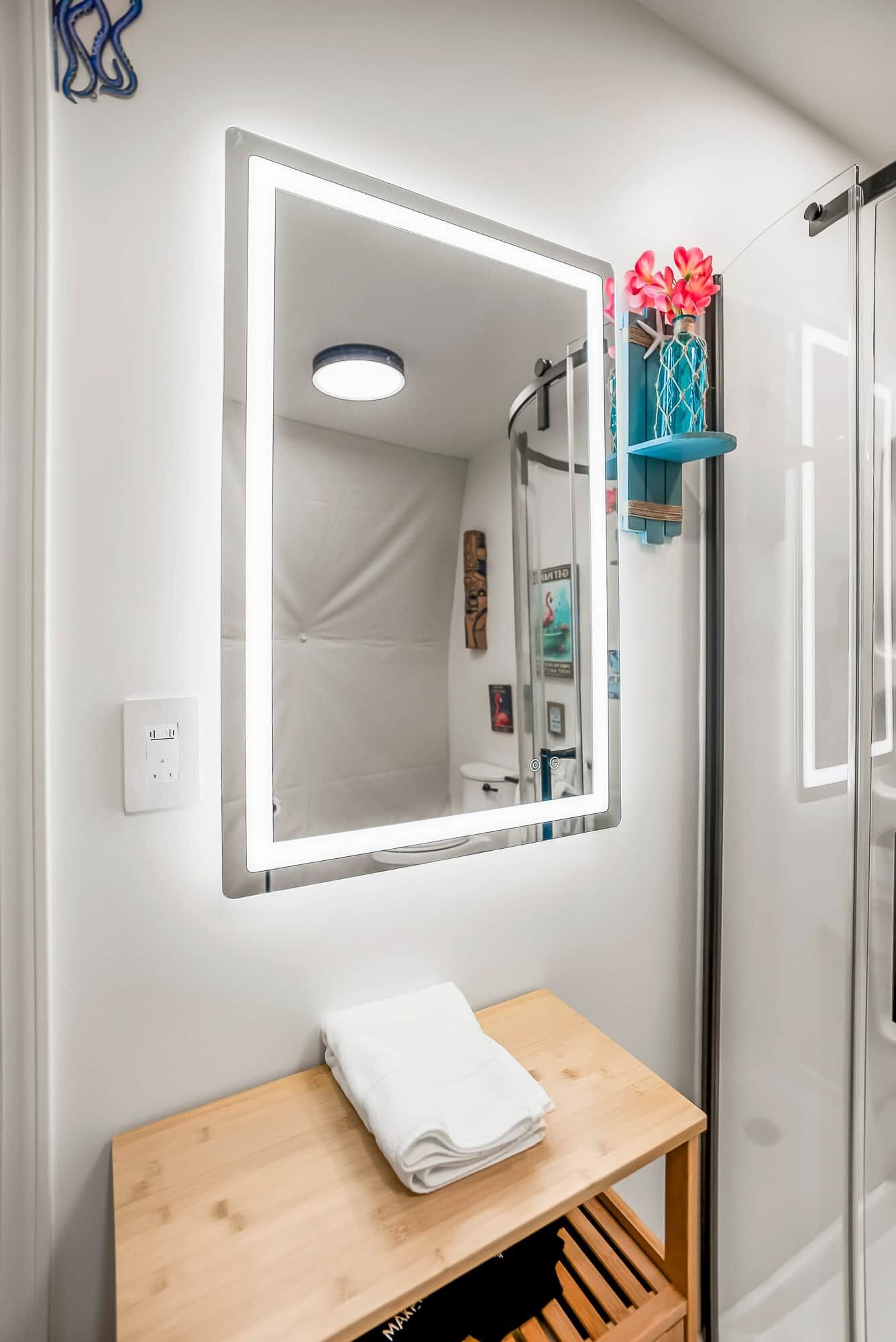 A bathroom with a mirror, a towel on a wooden shelf, a shower, and a wall-mounted light fixture.