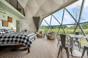 Ein Zelten-ähnliches Zimmer mit einem Bett, Stühlen, einem Tisch und Blick ins Grüne.
