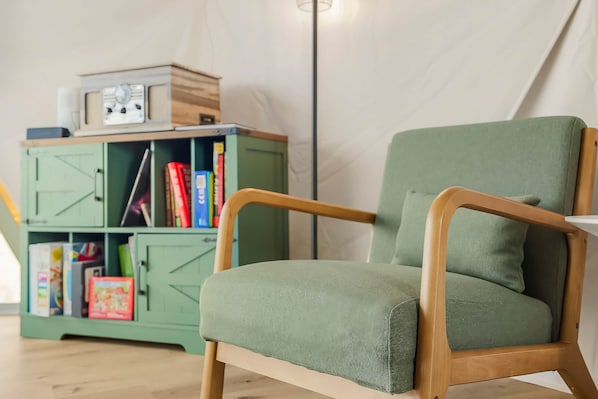 A green armchair with wooden armrests and a cushion, positioned in front of a green cabinet with books and a small radio.