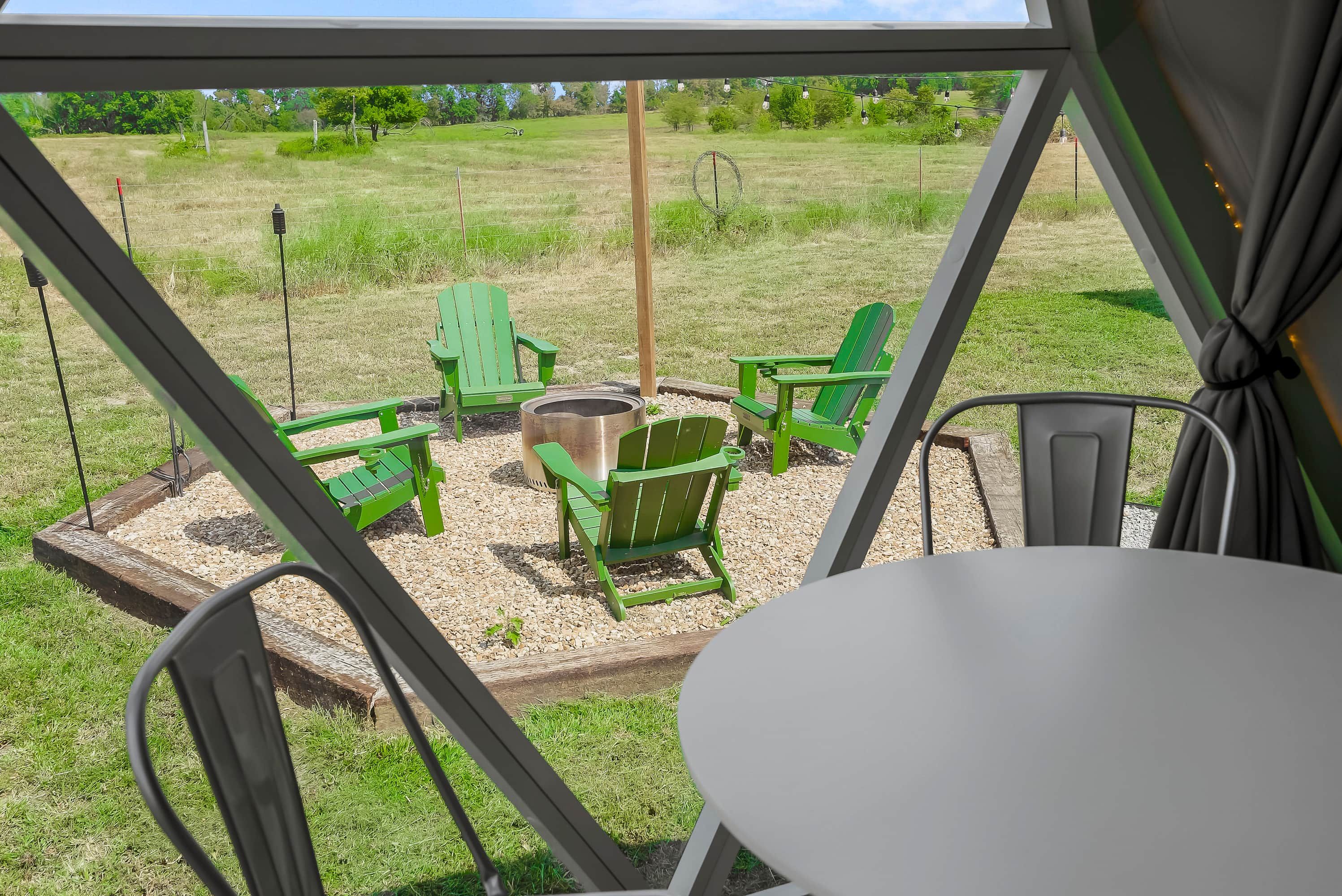 Ein überdachter Außenbereich mit Tisch und Stühlen, einem Feuerkorb und Blick auf ein saftiges Grasfeld.