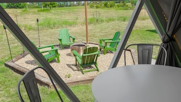 Ein überdachter Außenbereich mit Tisch und Stühlen, einem Feuerkorb und Blick auf ein saftiges Grasfeld.