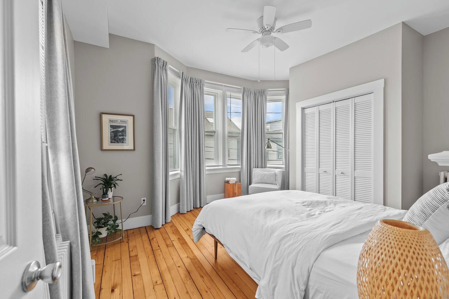 A bedroom with a bed, a ceiling fan, a bedside table with a lamp, a window with curtains, and a plant.