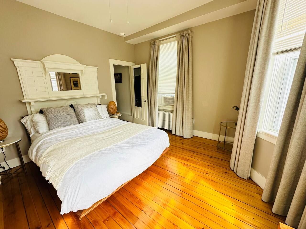 Une chambre à coucher avec un grand lit, une tête de lit blanche, une petite table et une fenêtre avec des rideaux.