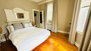 Une chambre à coucher avec un grand lit, une tête de lit blanche, une petite table et une fenêtre avec des rideaux.