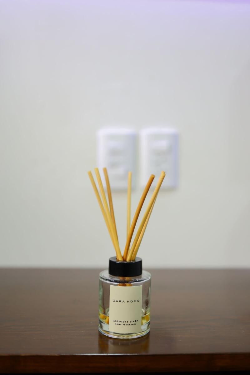 Une bouteille en verre transparent avec un bouchon noir, portant l’étiquette « ZARA HOME ABSOLUTE LINEN ROSE PERFUME », et contenant des bâtonnets parfumés disposés sur une surface en bois.