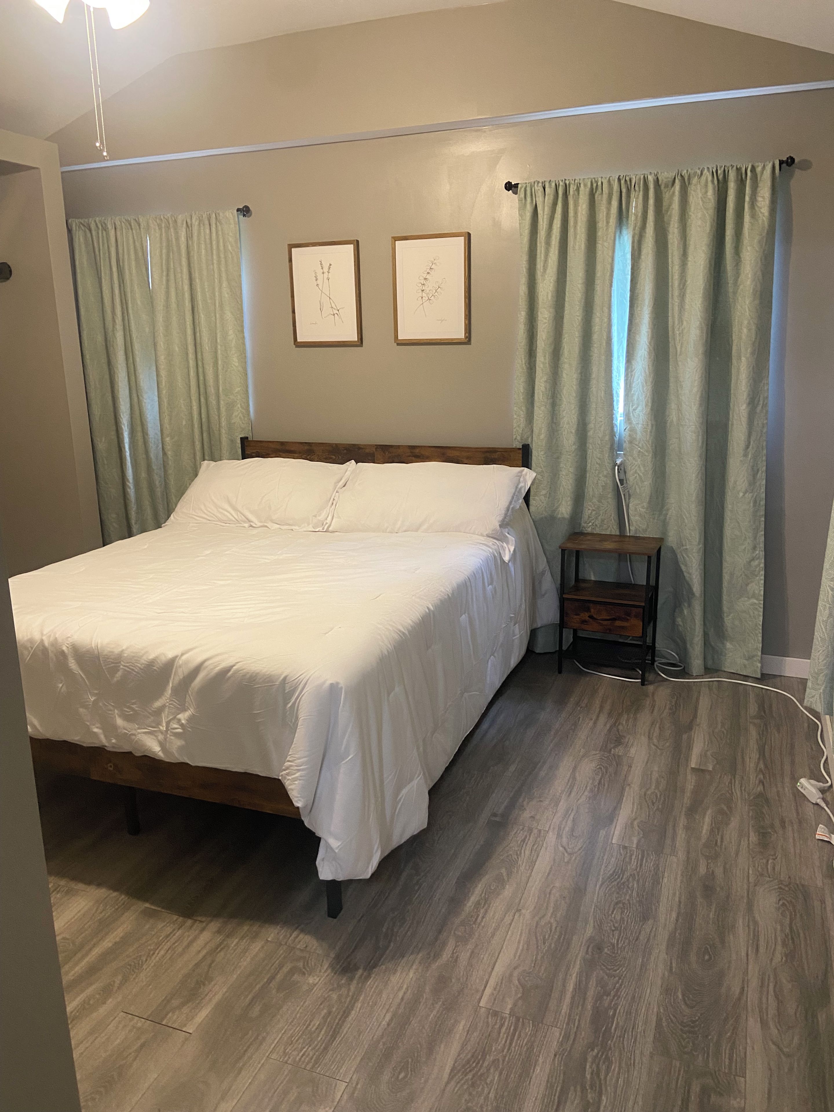 Un dormitorio con una cama de madera, cortinas verde claro y dos láminas botánicas enmarcadas en la pared.